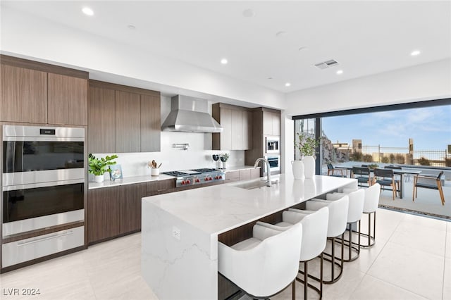 kitchen with sink, wall chimney exhaust hood, an island with sink, light stone counters, and stainless steel appliances