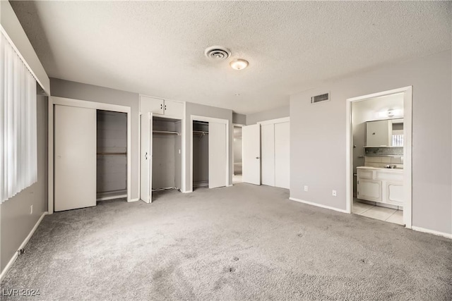 unfurnished bedroom with a textured ceiling, ensuite bathroom, light carpet, and two closets