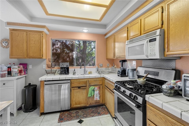 kitchen featuring stainless steel appliances, a tray ceiling, sink, light tile patterned floors, and tile countertops