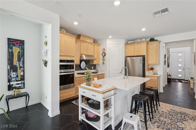 kitchen with light brown cabinets, a kitchen island with sink, dark tile patterned flooring, appliances with stainless steel finishes, and tasteful backsplash