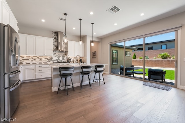 kitchen featuring stainless steel refrigerator with ice dispenser, wall chimney range hood, pendant lighting, a center island with sink, and white cabinetry