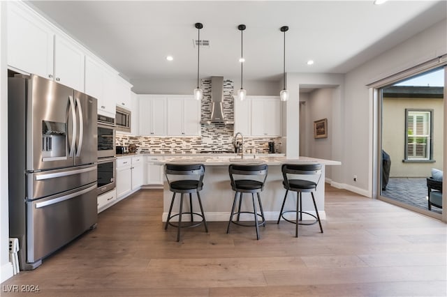 kitchen featuring a breakfast bar, wall chimney exhaust hood, a center island with sink, white cabinets, and appliances with stainless steel finishes