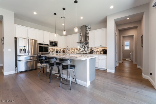 kitchen featuring wall chimney exhaust hood, stainless steel appliances, light hardwood / wood-style floors, a center island with sink, and white cabinets