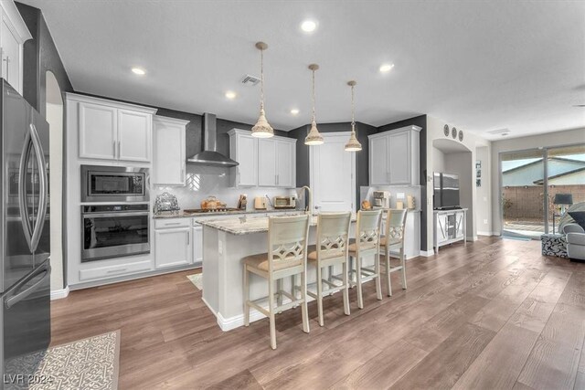 kitchen with wall chimney exhaust hood, white cabinetry, stainless steel appliances, an island with sink, and hanging light fixtures