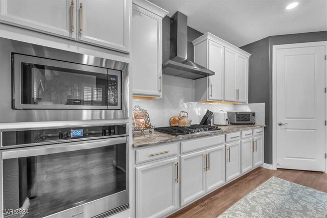 kitchen featuring light stone countertops, hardwood / wood-style floors, white cabinets, appliances with stainless steel finishes, and wall chimney exhaust hood