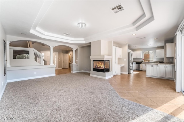 unfurnished living room featuring a multi sided fireplace, a tray ceiling, ornate columns, and light tile patterned flooring