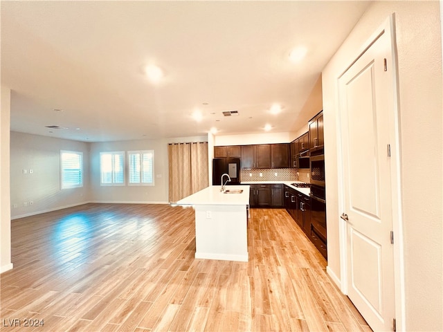 kitchen with backsplash, a kitchen island with sink, dark brown cabinets, black appliances, and light wood-type flooring