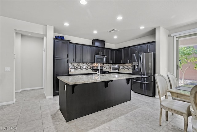 kitchen featuring light stone countertops, stainless steel appliances, an island with sink, a kitchen bar, and decorative backsplash