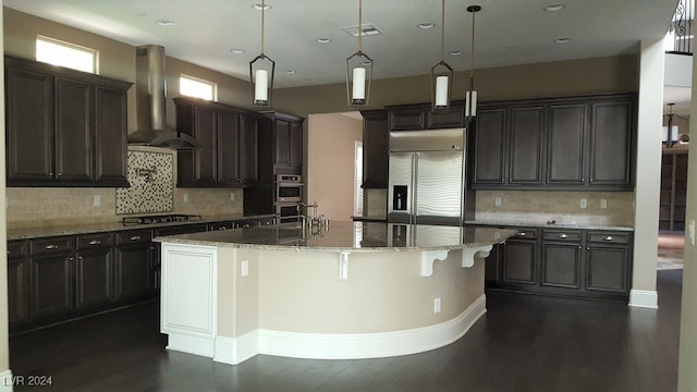 kitchen featuring wall chimney exhaust hood, light stone counters, an island with sink, and appliances with stainless steel finishes