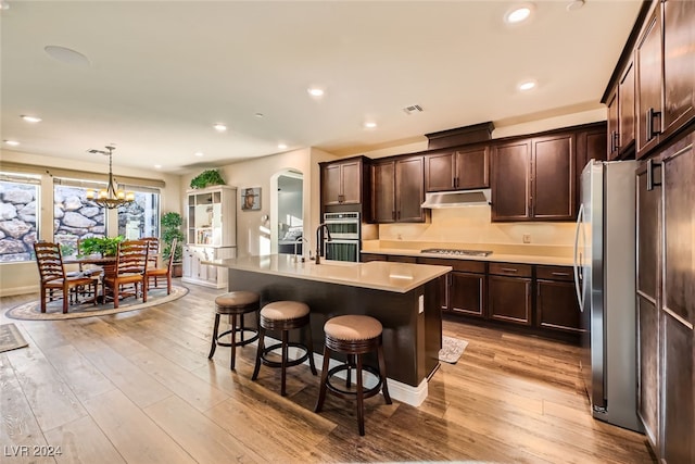 kitchen featuring light wood-type flooring, stainless steel appliances, decorative light fixtures, a center island with sink, and a chandelier