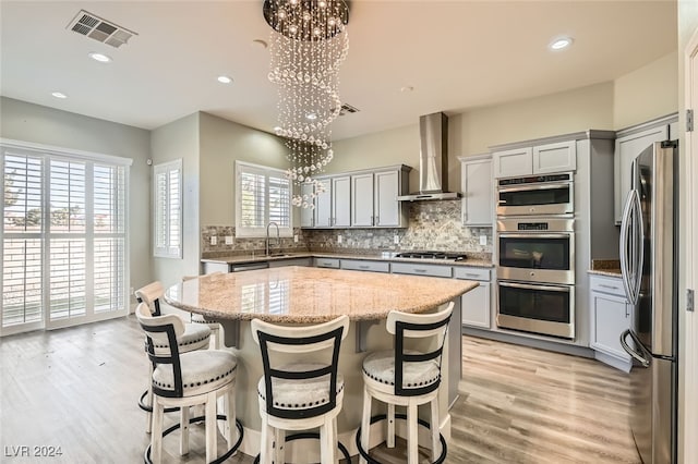 kitchen featuring wall chimney exhaust hood, light stone counters, pendant lighting, a kitchen island, and appliances with stainless steel finishes