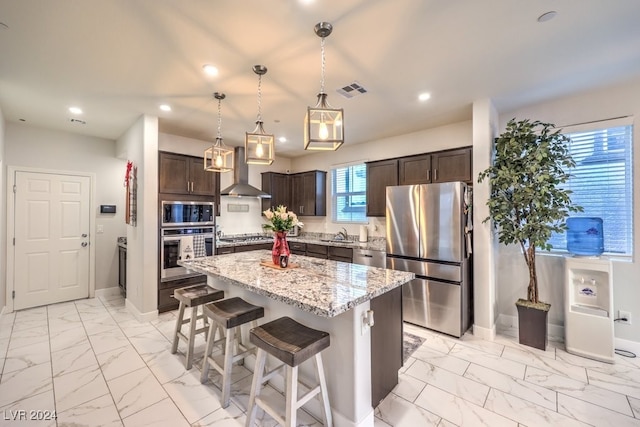 kitchen featuring appliances with stainless steel finishes, wall chimney exhaust hood, dark brown cabinets, decorative light fixtures, and a center island