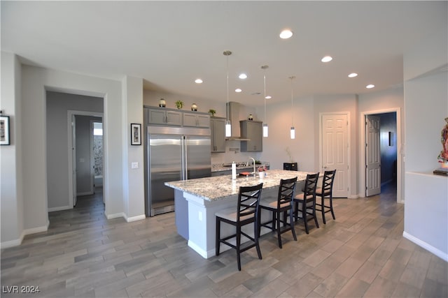 kitchen with hardwood / wood-style floors, built in refrigerator, light stone countertops, an island with sink, and decorative light fixtures