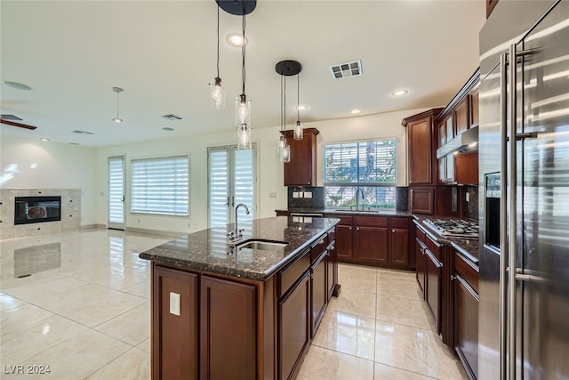 kitchen featuring dark stone counters, a center island with sink, sink, a premium fireplace, and appliances with stainless steel finishes