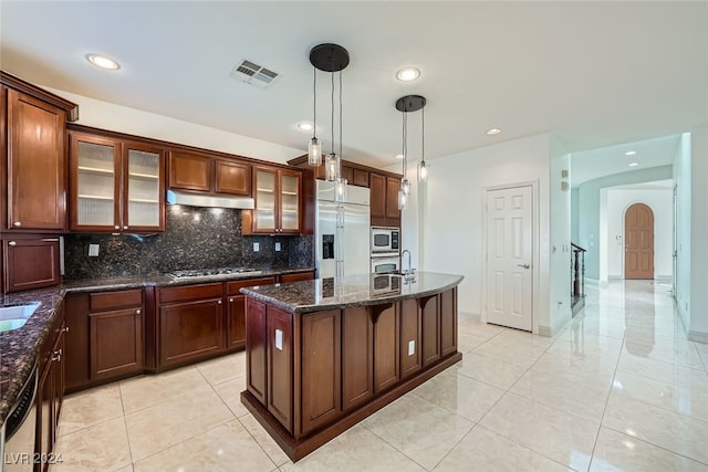 kitchen featuring tasteful backsplash, built in appliances, dark stone counters, decorative light fixtures, and a center island with sink