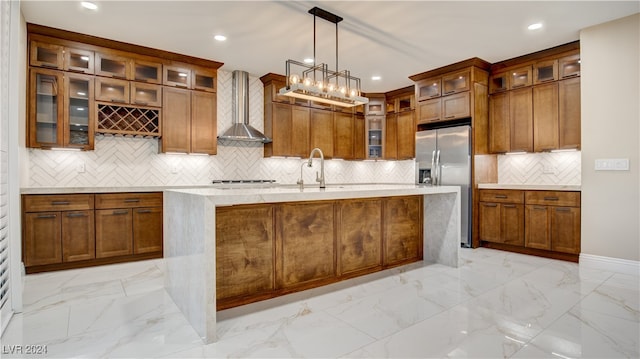 kitchen featuring pendant lighting, a center island with sink, wall chimney range hood, decorative backsplash, and stainless steel fridge