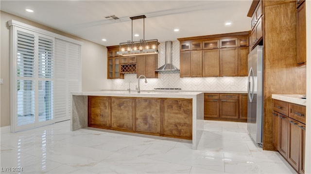 kitchen with decorative backsplash, stainless steel fridge, wall chimney exhaust hood, a kitchen island with sink, and decorative light fixtures