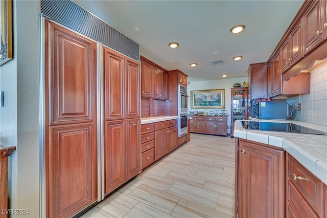 kitchen with black electric stovetop, tasteful backsplash, stainless steel double oven, paneled built in fridge, and tile counters