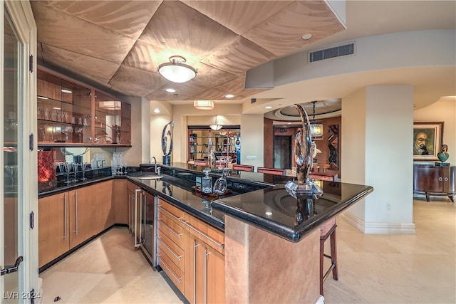 kitchen featuring kitchen peninsula, a kitchen bar, dark stone counters, beverage cooler, and sink