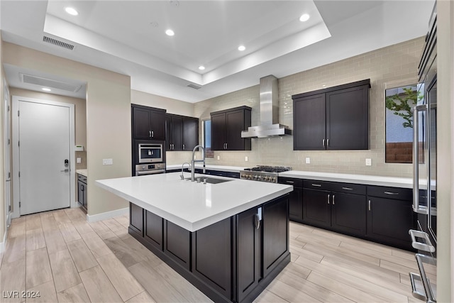 kitchen featuring sink, wall chimney range hood, a raised ceiling, a kitchen island with sink, and appliances with stainless steel finishes