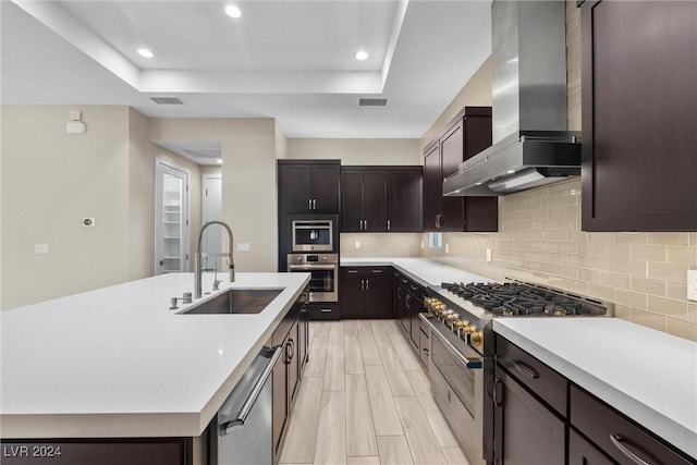 kitchen with stainless steel appliances, a raised ceiling, a kitchen island with sink, sink, and wall chimney range hood