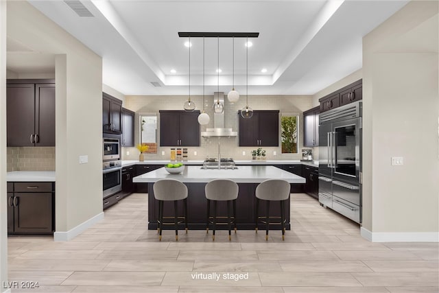 kitchen with dark brown cabinets, a raised ceiling, wall chimney range hood, a center island with sink, and stainless steel built in fridge