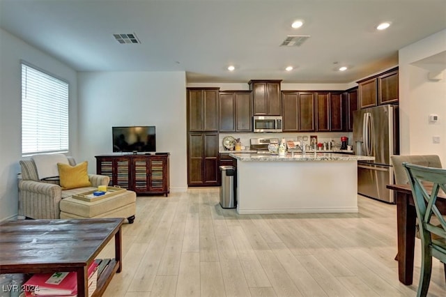 kitchen with dark brown cabinetry, light stone countertops, light hardwood / wood-style floors, a center island with sink, and appliances with stainless steel finishes