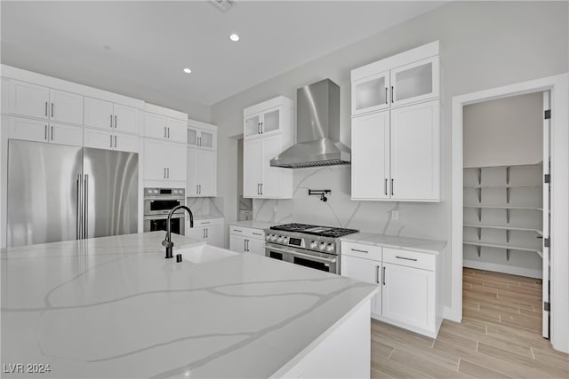 kitchen with light stone counters, premium appliances, sink, wall chimney range hood, and white cabinets