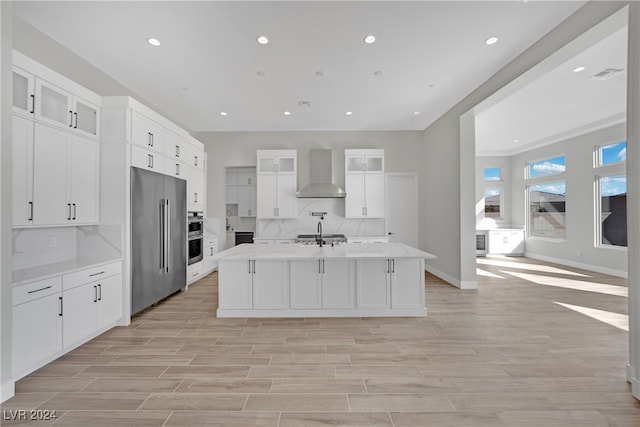 kitchen with appliances with stainless steel finishes, wall chimney range hood, white cabinets, light hardwood / wood-style floors, and an island with sink