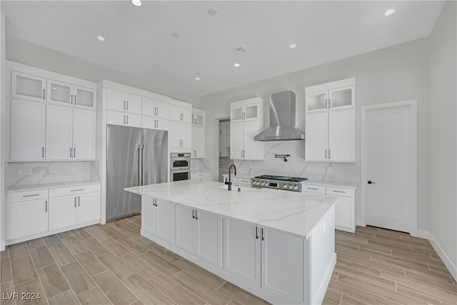 kitchen with light stone counters, wall chimney exhaust hood, stainless steel appliances, a kitchen island with sink, and white cabinets