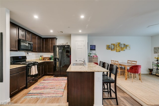 kitchen featuring light stone counters, an island with sink, a breakfast bar area, black appliances, and light wood-type flooring