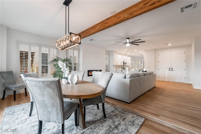 dining room featuring beam ceiling, ceiling fan, a large fireplace, and light wood-type flooring