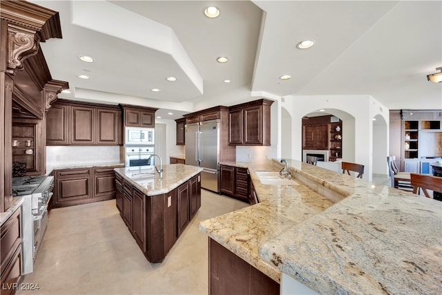 kitchen with tasteful backsplash, light stone counters, sink, built in appliances, and a center island with sink