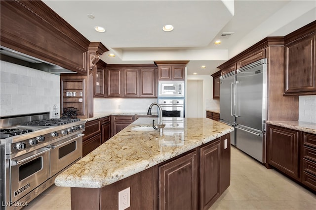 kitchen with sink, decorative backsplash, light stone counters, built in appliances, and a kitchen island with sink