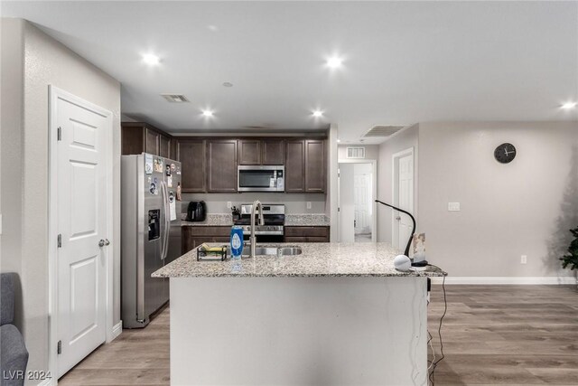 kitchen with light stone counters, dark brown cabinetry, stainless steel appliances, visible vents, and a center island with sink