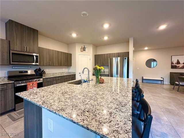 kitchen featuring dark brown cabinetry, a sink, appliances with stainless steel finishes, a large island, and light stone countertops