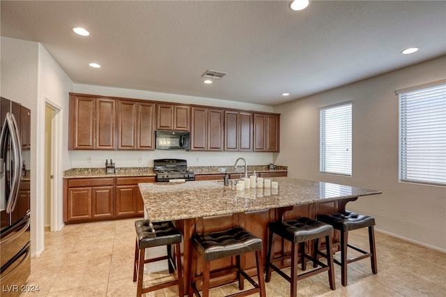 kitchen featuring a breakfast bar, black appliances, an island with sink, light tile patterned flooring, and light stone counters