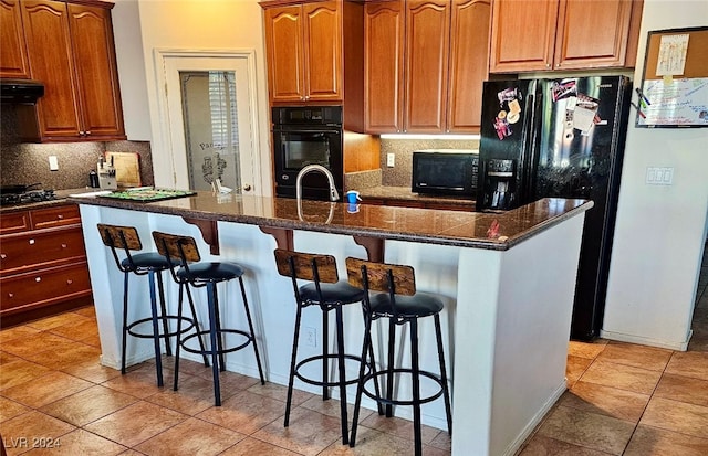 kitchen featuring a kitchen breakfast bar, dark stone counters, a kitchen island with sink, exhaust hood, and black appliances