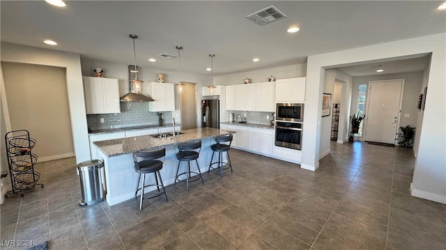 kitchen featuring stainless steel appliances, wall chimney range hood, dark stone countertops, white cabinets, and an island with sink