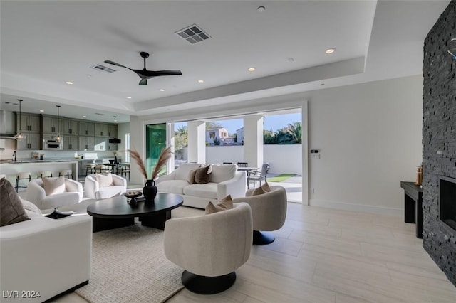 living room featuring a raised ceiling, a stone fireplace, ceiling fan, and sink