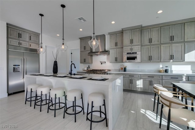 kitchen with wall chimney range hood, an island with sink, decorative light fixtures, a kitchen bar, and appliances with stainless steel finishes