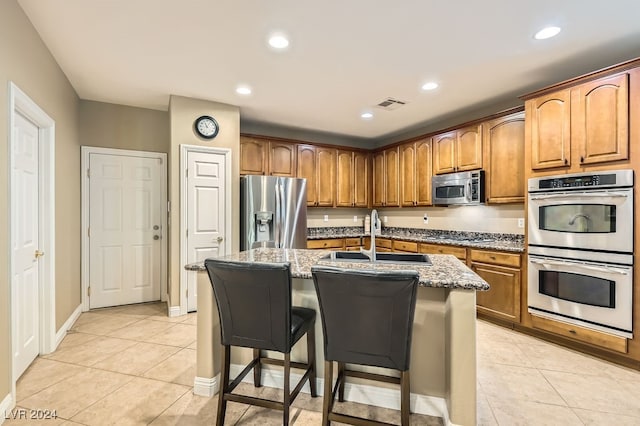 kitchen with sink, stainless steel appliances, light stone counters, an island with sink, and a breakfast bar area