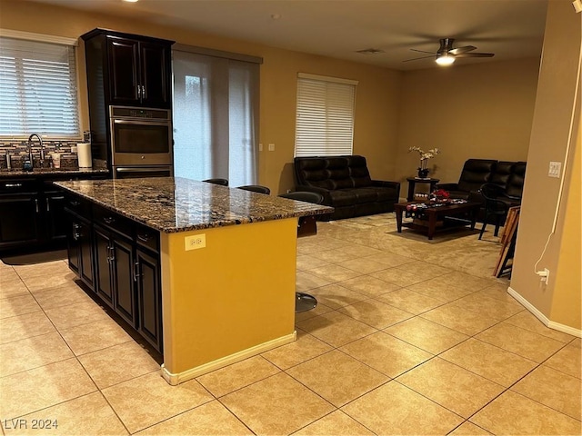 kitchen featuring dark stone counters, stainless steel double oven, ceiling fan, a center island, and light tile patterned flooring