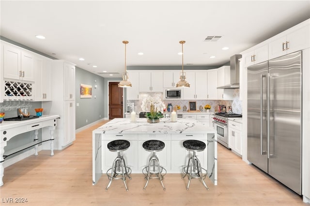 kitchen with white cabinetry, wall chimney exhaust hood, built in appliances, decorative light fixtures, and a center island with sink