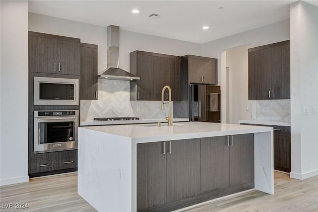 kitchen featuring decorative backsplash, wall chimney exhaust hood, an island with sink, and appliances with stainless steel finishes