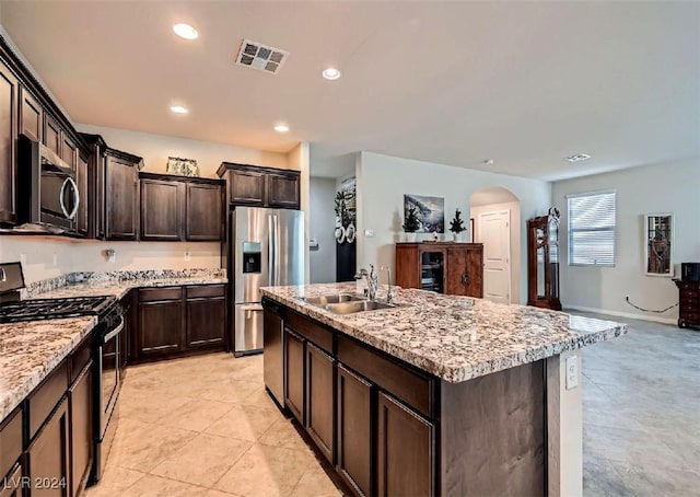 kitchen with a center island with sink, sink, dark brown cabinets, light stone counters, and stainless steel appliances