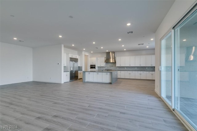 kitchen with wall chimney exhaust hood, appliances with stainless steel finishes, a center island with sink, white cabinets, and light wood-type flooring