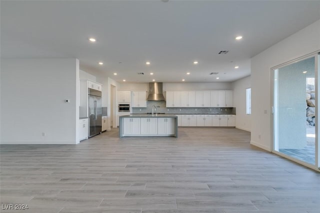 kitchen featuring a kitchen island with sink, wall chimney range hood, sink, appliances with stainless steel finishes, and white cabinetry