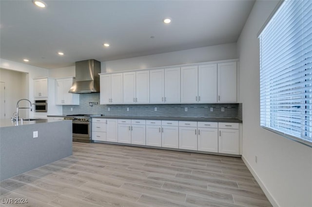 kitchen with white cabinets, wall chimney range hood, sink, light hardwood / wood-style flooring, and stainless steel appliances