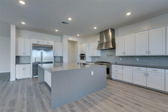 kitchen with white cabinetry, wall chimney range hood, premium appliances, light hardwood / wood-style flooring, and a center island with sink